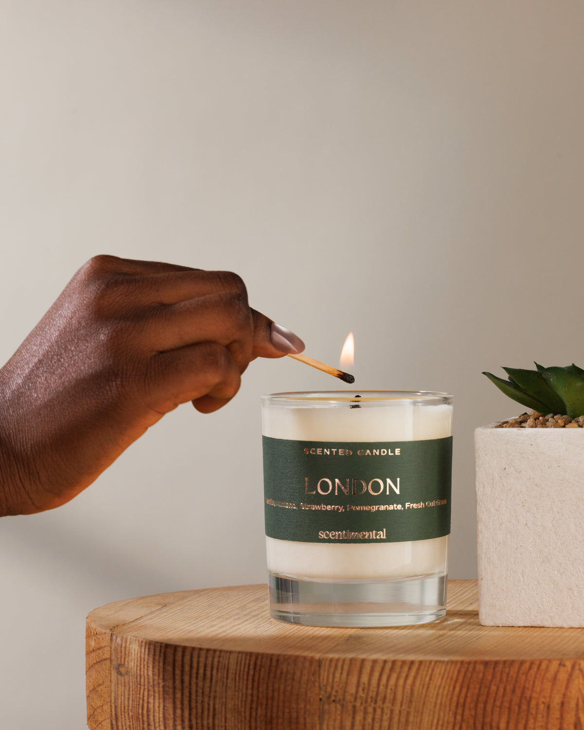 A hand holds a lit match above a scented candle in a clear glass jar with a dark green label reading 'LONDON' and 'scented candles' at the top, set on a wooden surface with a small potted succulent beside it.